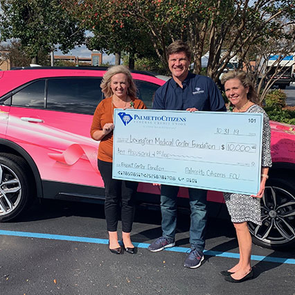 Two women and man standing next to pink car holding large check